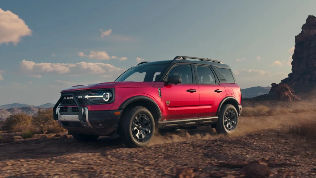 Red 2025 Ford Bronco Sport® driving on rocky terrain under a blue sky near desert cliffs.