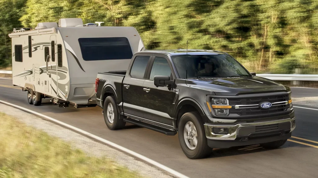 Black 2025 Ford F-150® towing a travel trailer on a highway surrounded by trees.