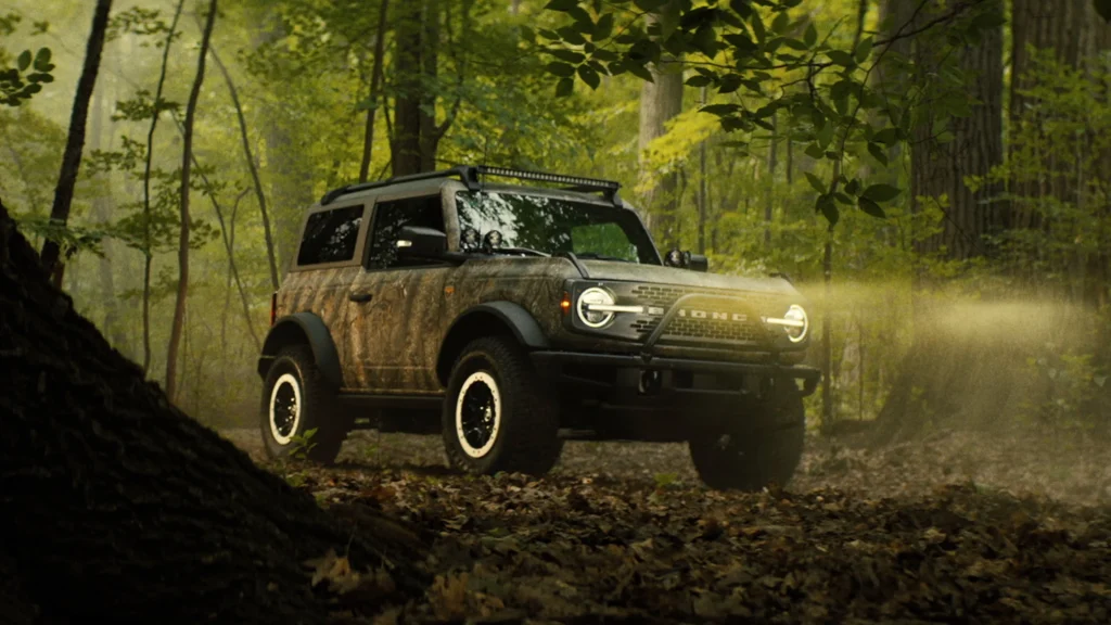 2025 Ford Bronco® in camouflage wrap with headlights on driving through a wooded trail at dusk.