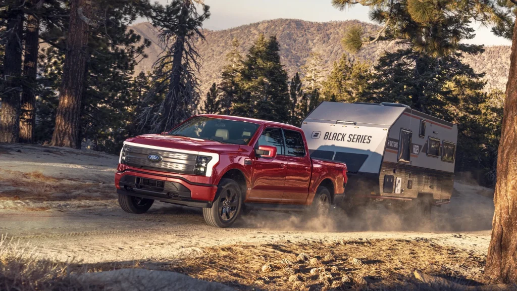 A red 2024 Ford F-150 Lightning electric truck towing a Black Series camper through a rugged forest trail, highlighting its towing power and off-road capability.