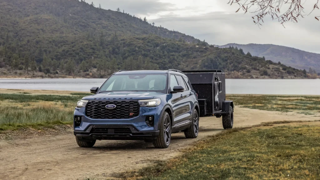 A blue 2025 Ford Explorer ST SUV towing a black utility trailer near a lake, emphasizing its towing performance and athletic design in a scenic outdoor setting.