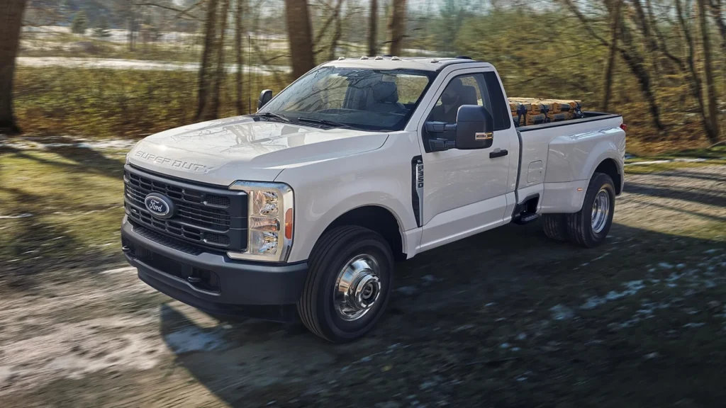 White 2025 Ford Super Duty truck hauling lumber through wooded dirt road with sunlight reflection.